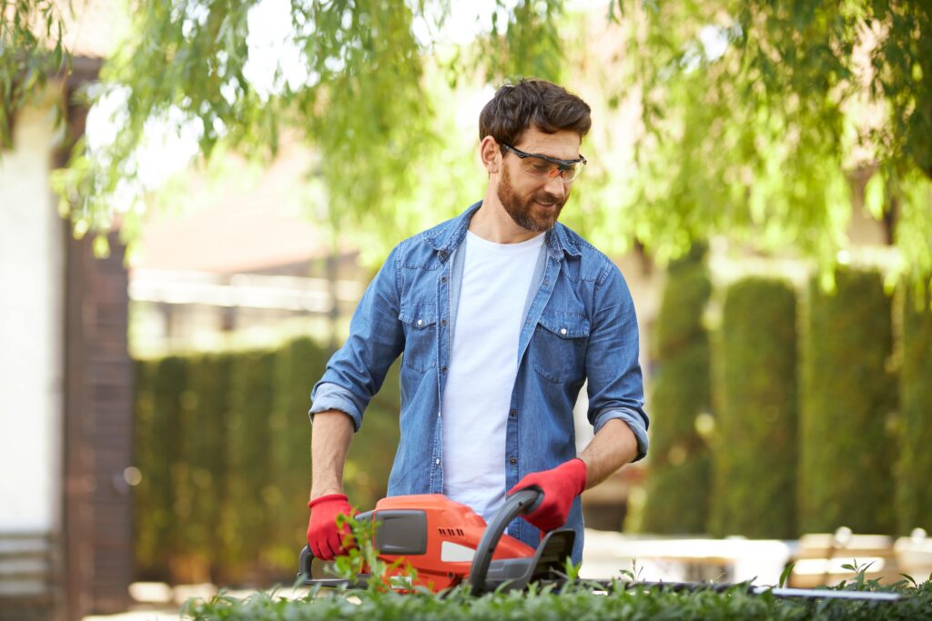 smiling-brunet-male-gardener-cutting-overgrown-bush-by-hedge-trimmer-sunny-day-front-view-hard-min
